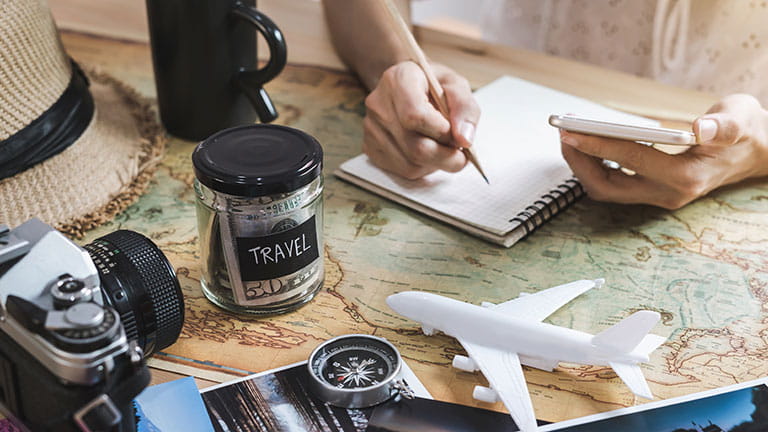 Person surrounded by travel-themed items with jar of money that says 